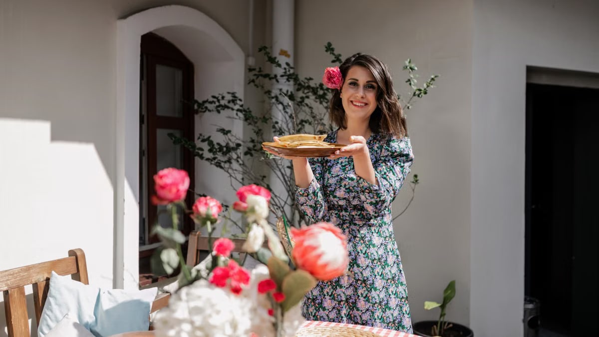 A woman in a floral dress holds plates at an outdoor garden party table with flowers.